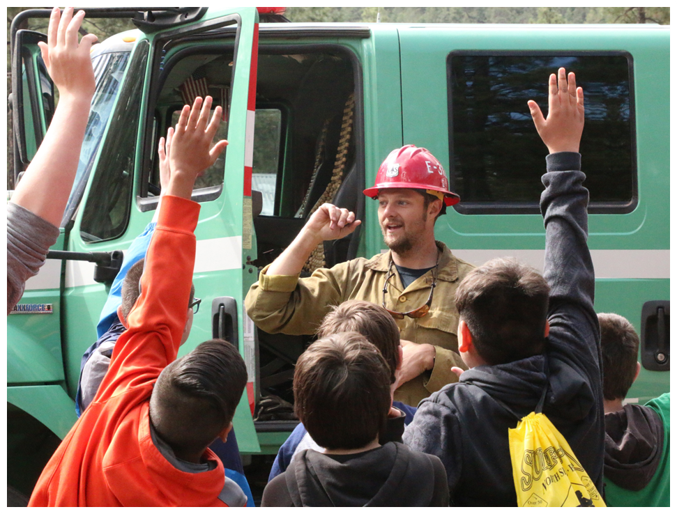 A firefighter stands in front of an eager to learn group of young people who have their hands raised.