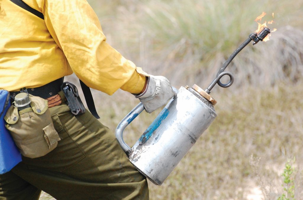 A fire fighter with a large pack, helmet & yellow jacket works on a fire.