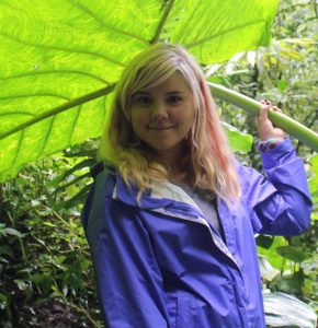 Anna is a woman with long blond hair standing under a big leaf and wearing a purple coat.