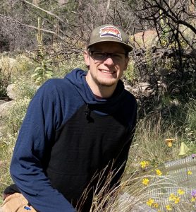 Ethan is a man crouching in a field of flowers, wearing a tan cap and navy shirt.