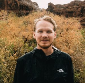Will is a man with short blond hair standing in front of a flowering shrub and sandstone cliffs. He is wearing a black shirt.