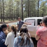 A group of children stand by a truck showing a map. A male teacher is pointing at the map.