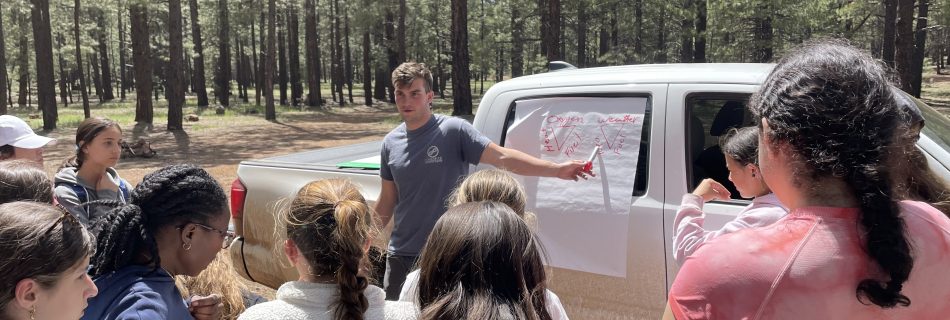 A group of children stand by a truck showing a map. A male teacher is pointing at the map.