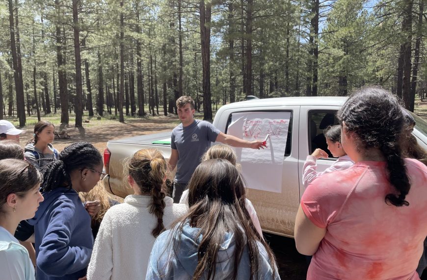 A group of children stand by a truck showing a map. A male teacher is pointing at the map.