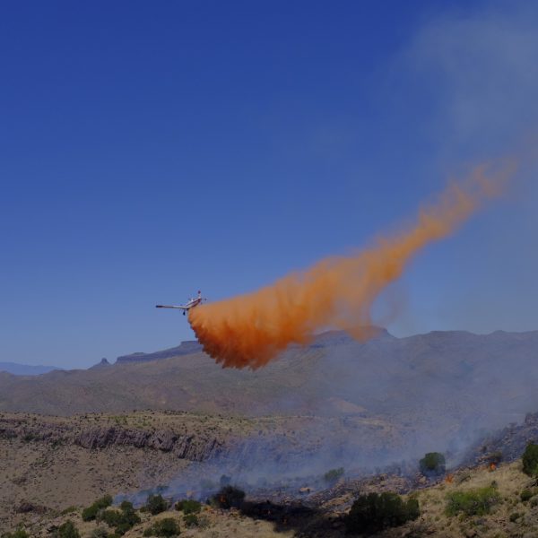 Blue skies, an airplane with redish orange fire retardant trailing behind. Smoky desert landscape below. A plane drops retardant on 2018 Sombrero Fire. Photo by BLM Arizona.