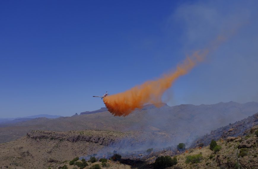 Blue skies, an airplane with redish orange fire retardant trailing behind. Smoky desert landscape below. A plane drops retardant on 2018 Sombrero Fire. Photo by BLM Arizona.